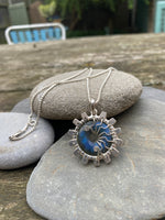 Silver cog necklace with a blue labradorite stone in the centre. On a silver chain. A background of stacked pebbles